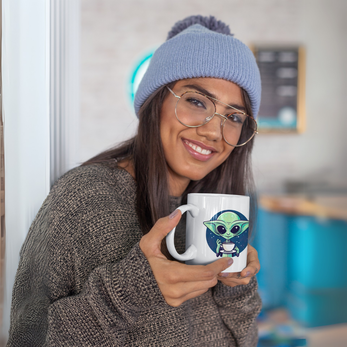 A young woman wearing a cap and glasses smiles friendly and presents her coffee mug, which features a cute alien.