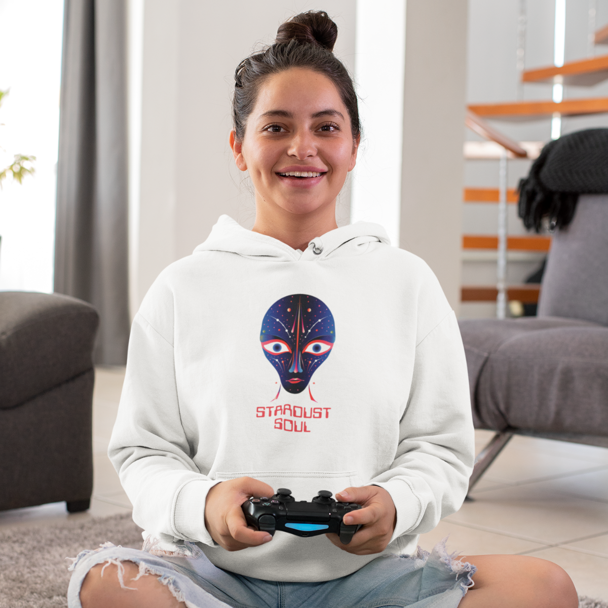 A girl is sitting smiling, holding a game controller, and wearing a white hoodie with an alien headprint on it. 