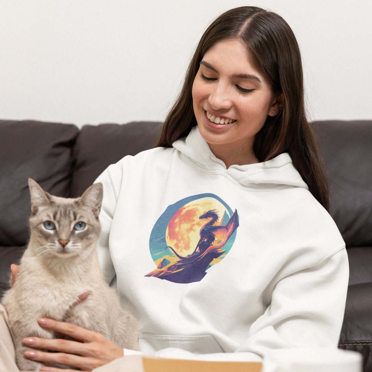 A young woman is sitting in the living room, holding her cat and wearing a white hoodie with a colorful dragon print.