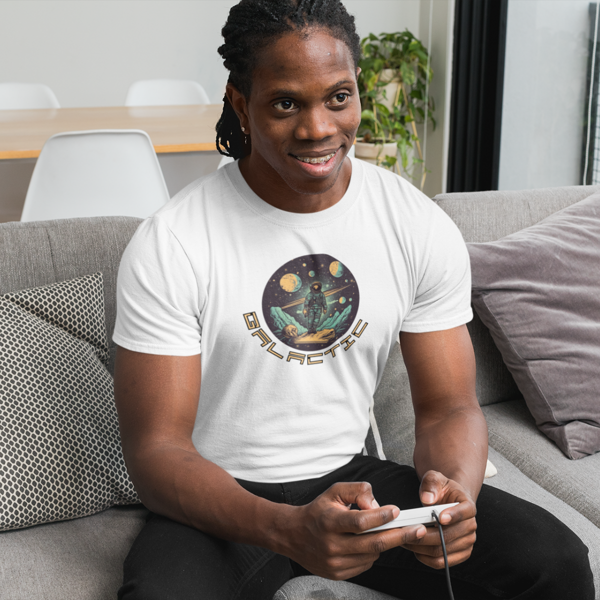 A man is sitting on a couch in a living room, holding a video game controller. He is wearing a t-shirt with an astronaut print.