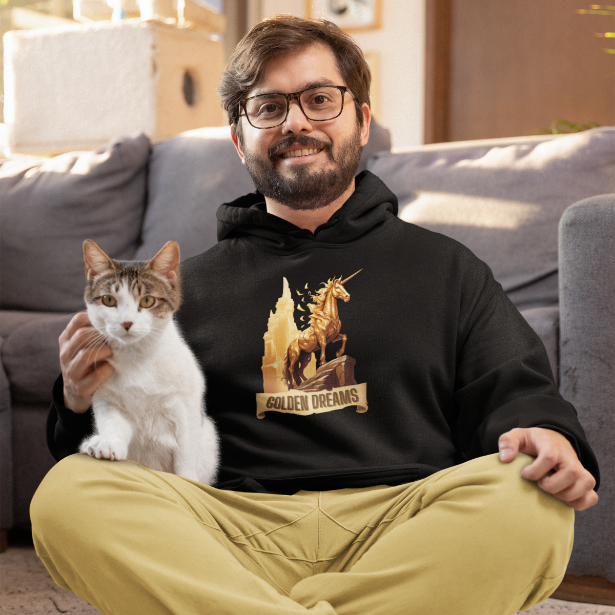 A man sits in the living room with his cat in his hand. He is wearing a black hoodie with a unicorn motif.