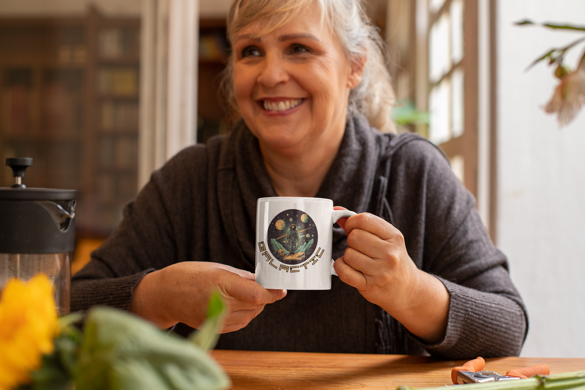 an elderly woman sits at the living room table laughing and holding a cup with an astronaut design on it.