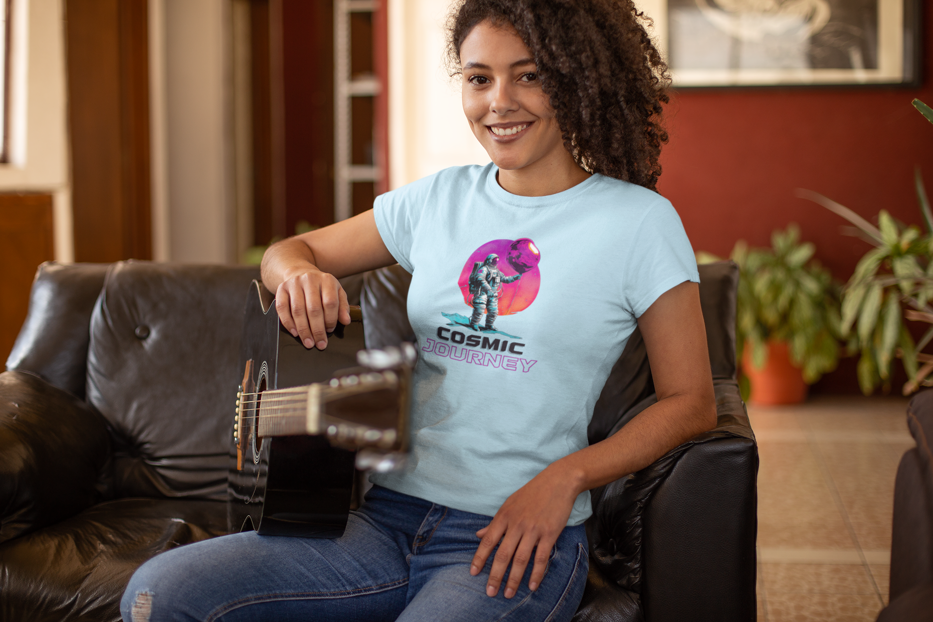 A woman with curly hair sits on a sofa and holds a guitar in her hand. She is wearing a light blue shirt with an astronaut print on it.