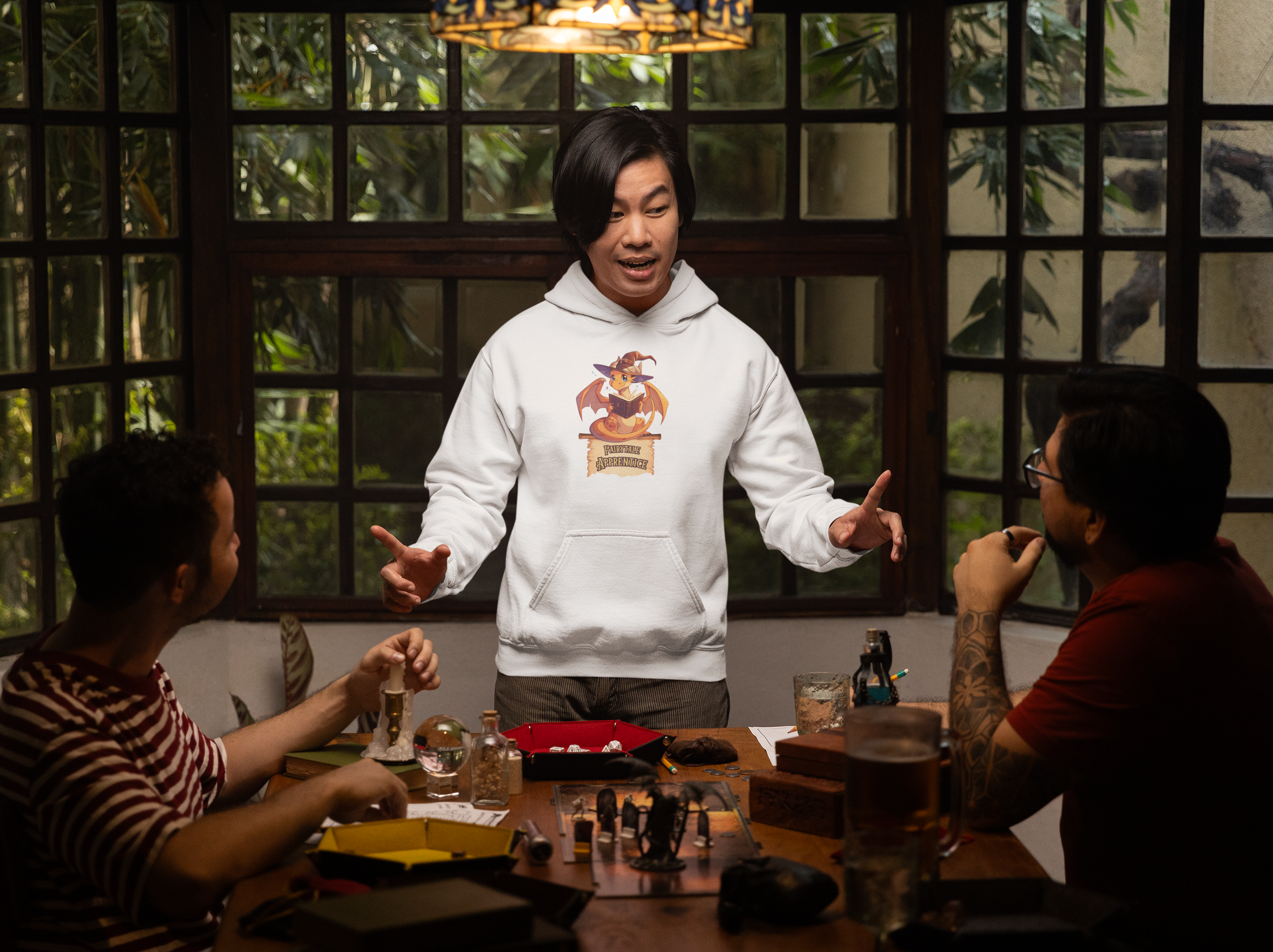 a young man stands in a living room talking to his friends, wearing a hoodie with a dragon on it