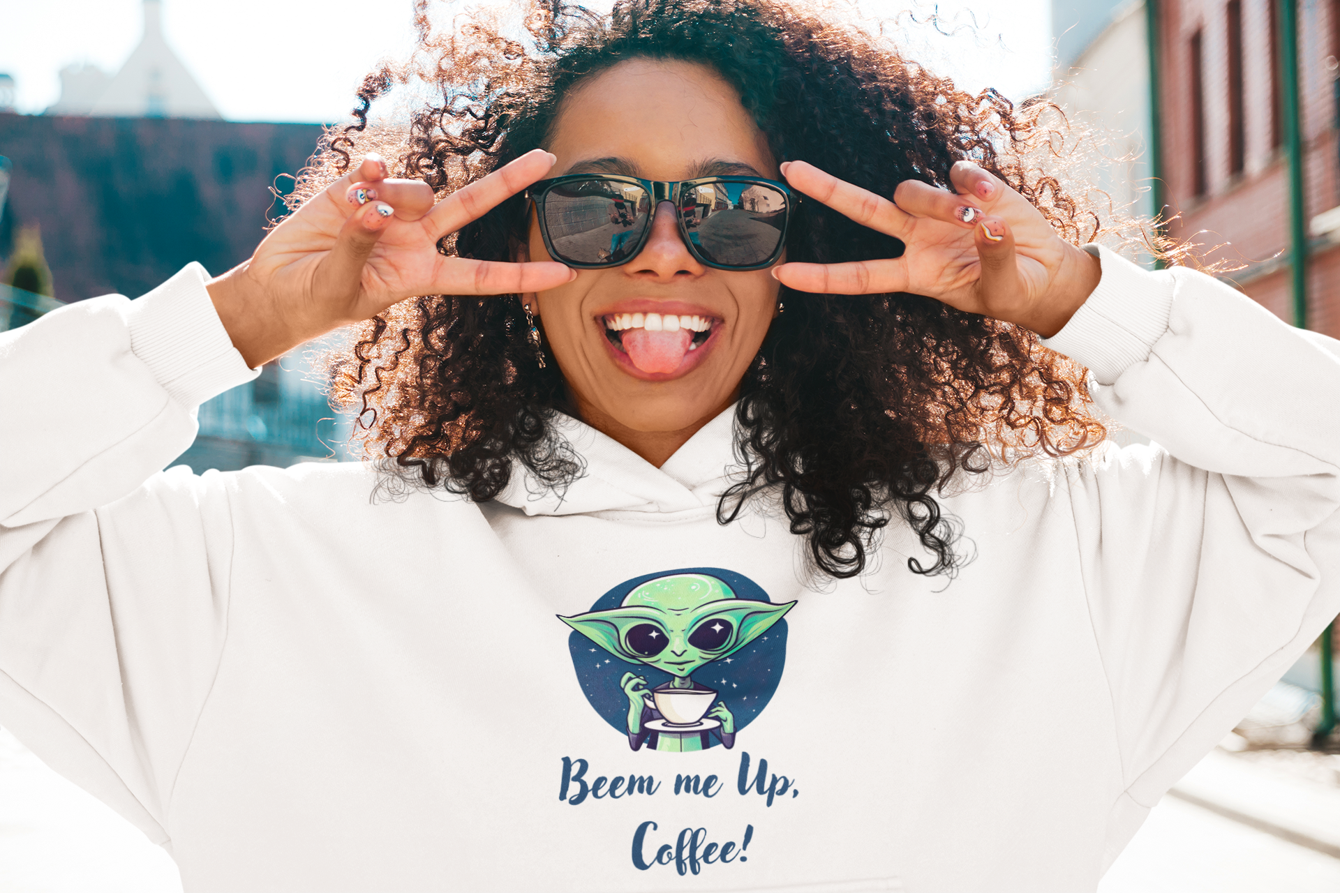 A young woman is wearing sunglasses and laughing at the camera. She is wearing a hoodie with an alien motif on it.