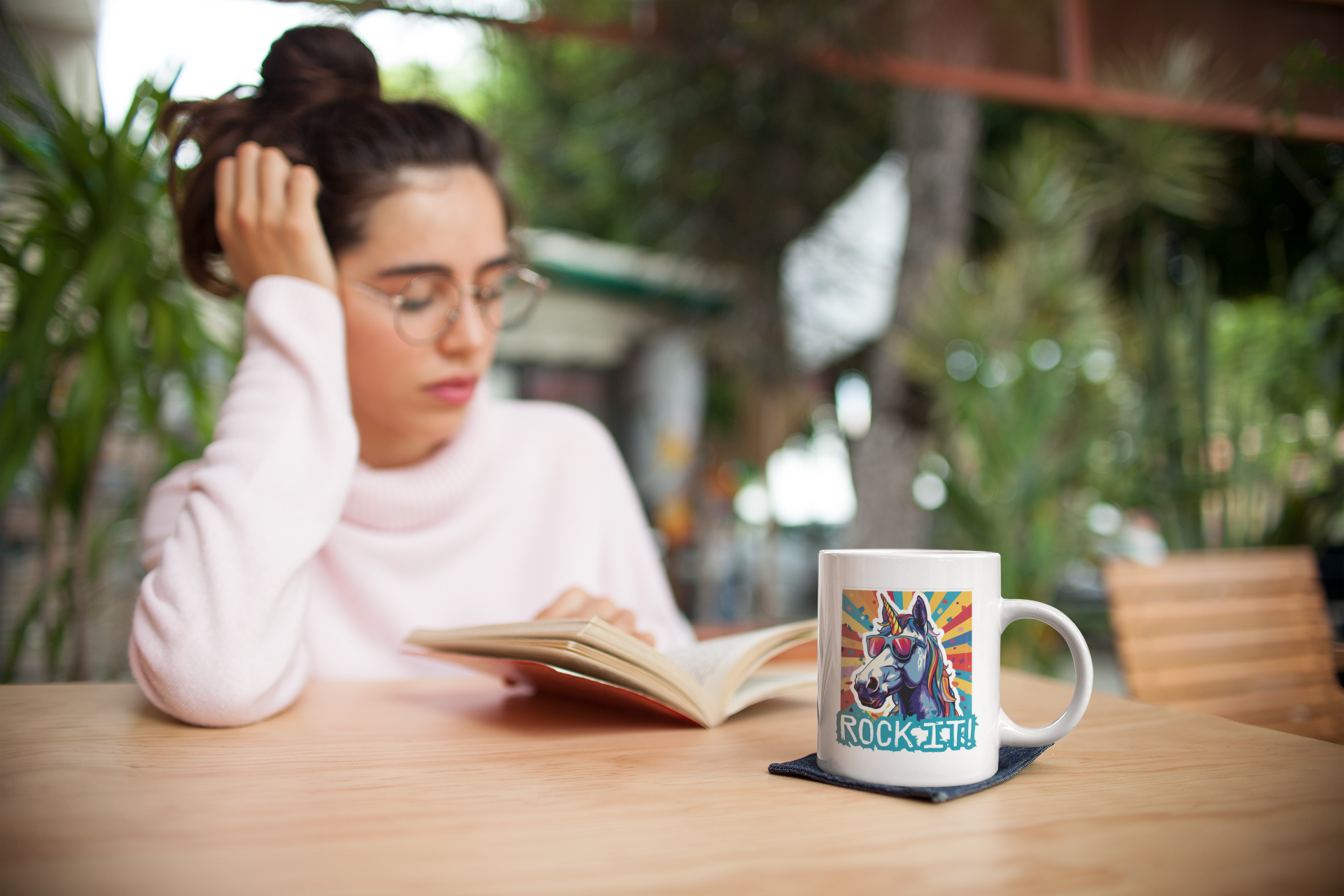 A young woman is sitting at a table reading a book, in front of her is a white coffee cup with a colourful unicorn motif on it.