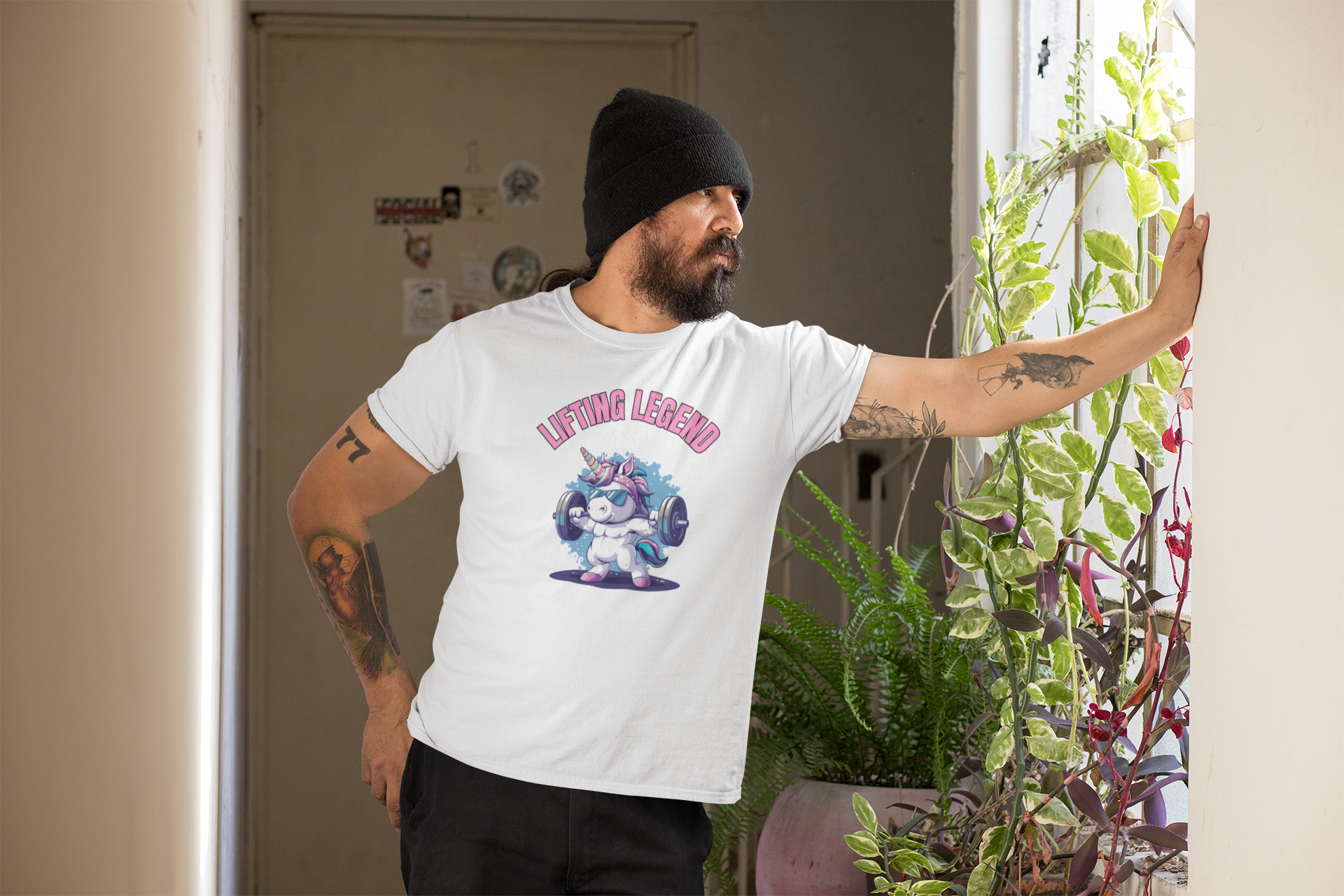 A man with a beard and cap leans against a wall and looks into the distance. He is wearing a white shirt with a sporty unicorn motif on it.